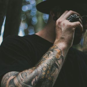 Tattooed man adjusting sunglasses amidst bokeh effect in forest setting.