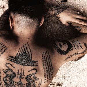 A tattooed man with intricate designs lying on a sandy beach, showcasing body art.