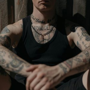 Close-up of a tattooed person in a black tank top sitting against a corrugated wall in moody lighting.