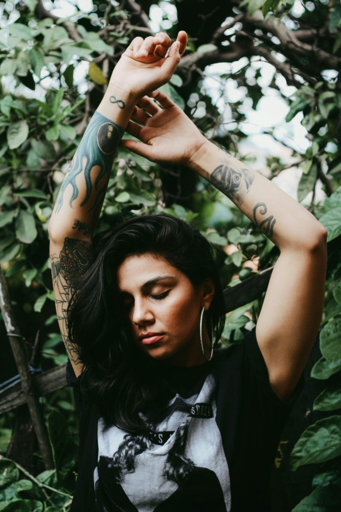 Portrait of a woman with tattoos posing outdoors, surrounded by lush greenery.