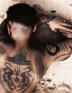 A tattooed man with intricate designs lying on a sandy beach, showcasing body art.