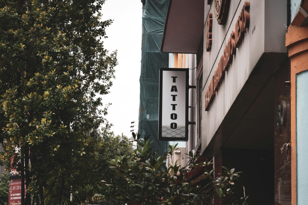 Street view of a tattoo shop signboard amidst urban buildings and greenery.